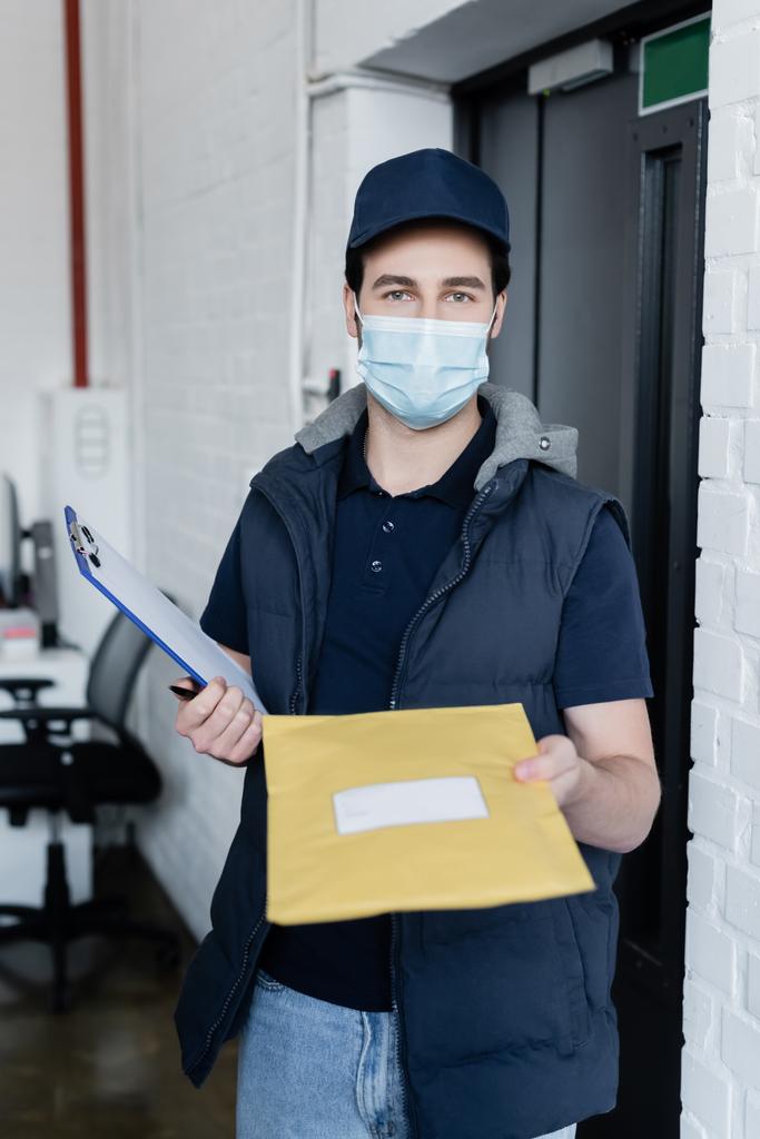 Courier in medical mask holding clipboard and parcel while looking at camera in office  - Фото, изображение
