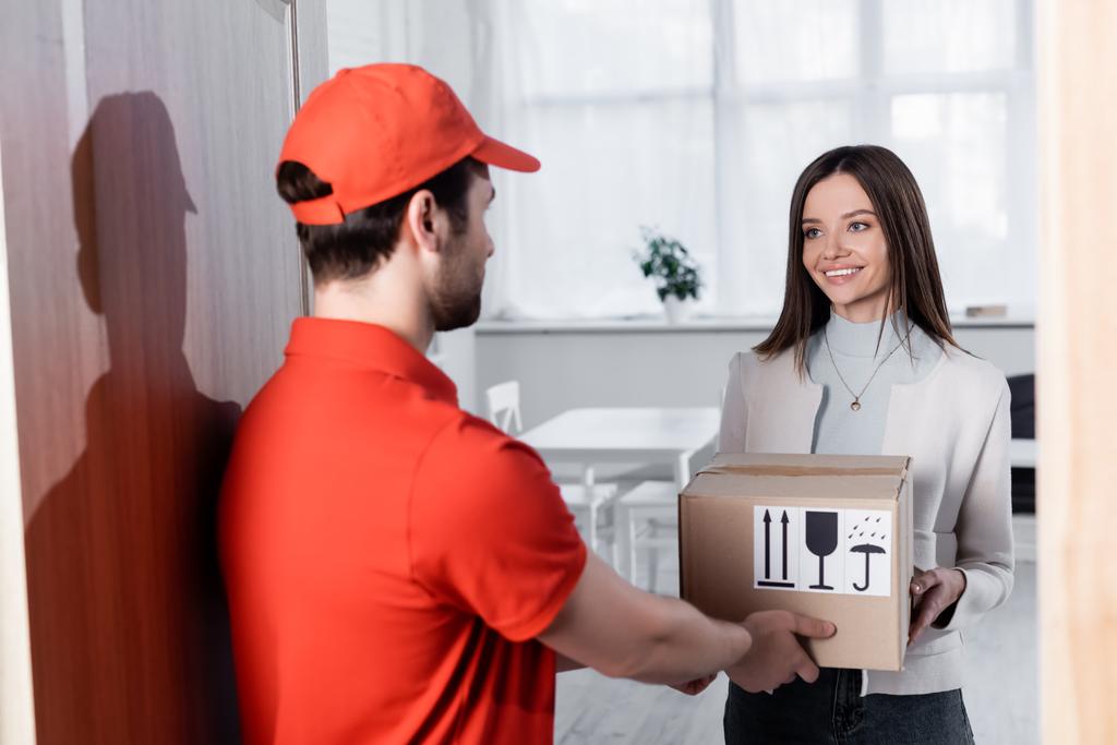 Blurred courier giving carton box to smiling woman in hallway  - Fotografie, Obrázek