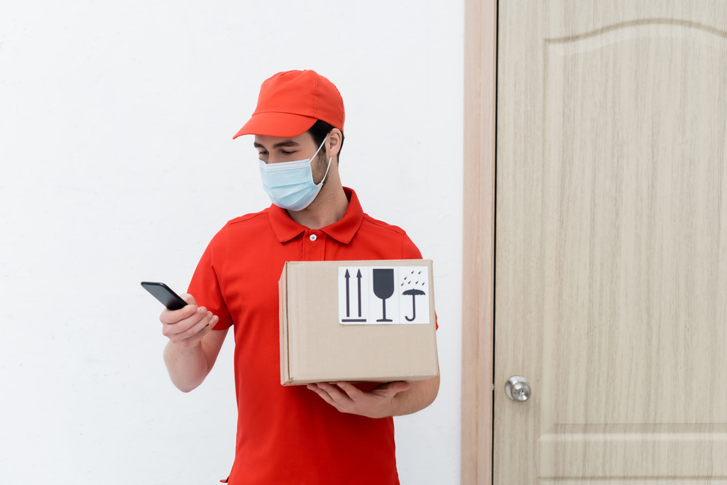 Delivery man in medical mask using smartphone and holding carton box in hallway  - Foto, imagen