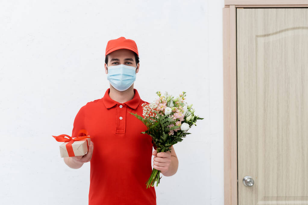Delivery man in medical mask holding bouquet and present in hallway  - Photo, image