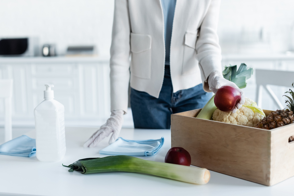 Cropped view of woman in latex gloves taking apple from box near rags and antiseptic at home  - Фото, изображение