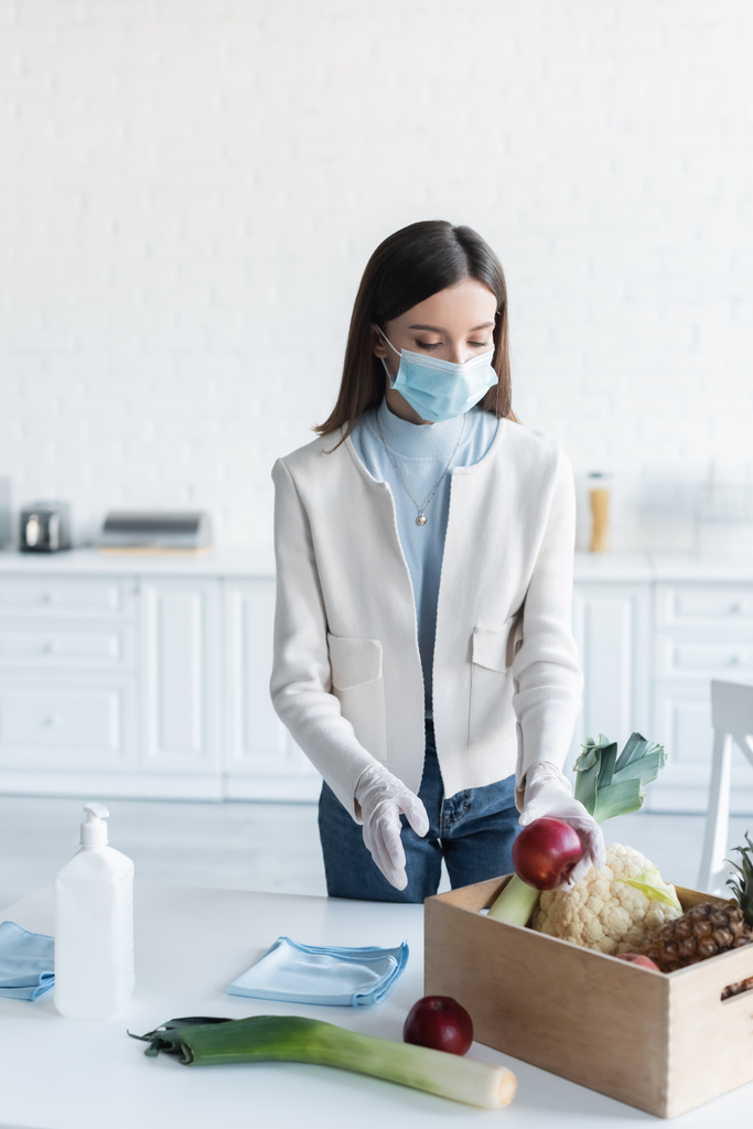 Woman in medical mask and latex gloves taking food from box near antiseptic in kitchen  - Фото, изображение