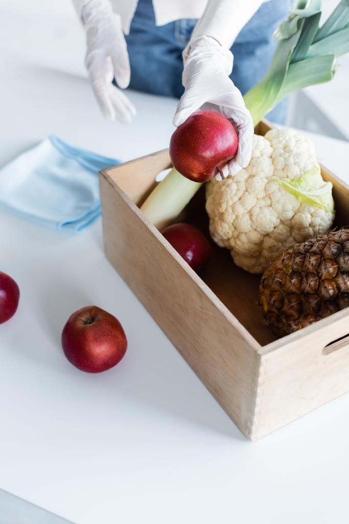Cropped view of woman in latex gloves holding apple near wooden box with fresh food and rag at home  - Photo, Image