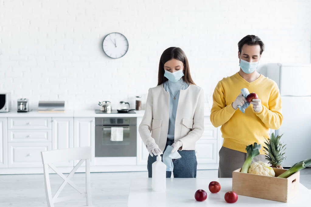 Couple in medical masks and latex gloves cleaning fresh food with antiseptic in kitchen  - Foto, imagen