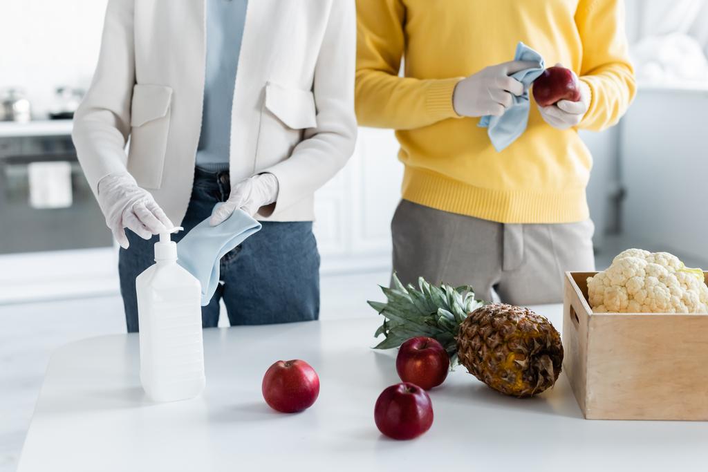 Cropped view of couple in latex gloves cleaning fresh food with antiseptic near box in kitchen  - Foto, imagen