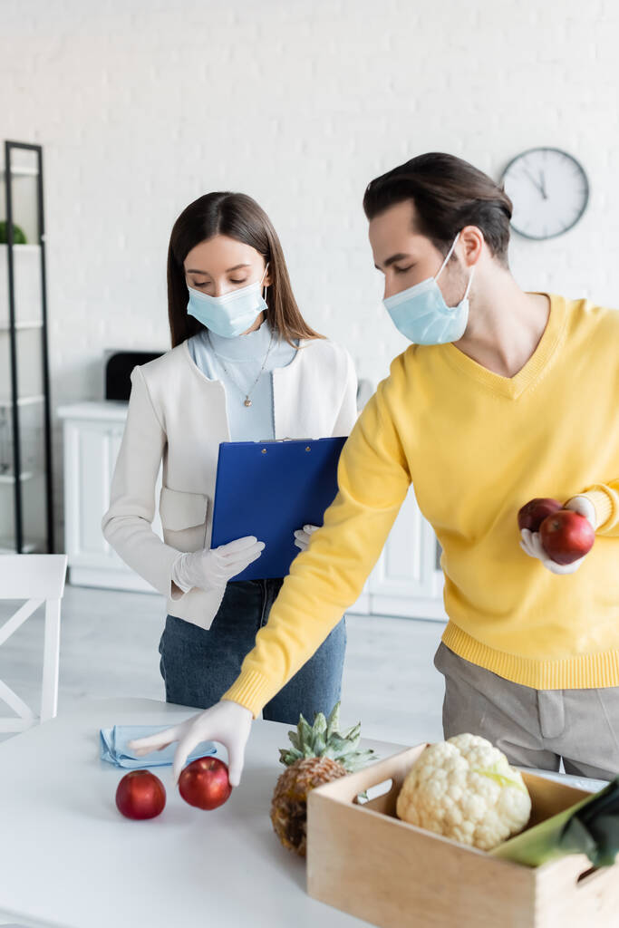 Man in medical mask holding apples near girlfriend in latex gloves holding clipboard in kitchen  - Foto, Bild