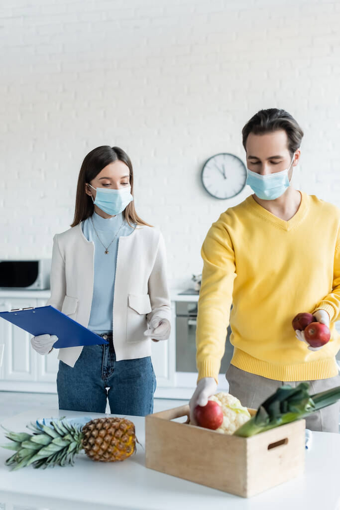Man in latex gloves and medical mask holding apples near girlfriend with clipboard in kitchen  - Fotografie, Obrázek