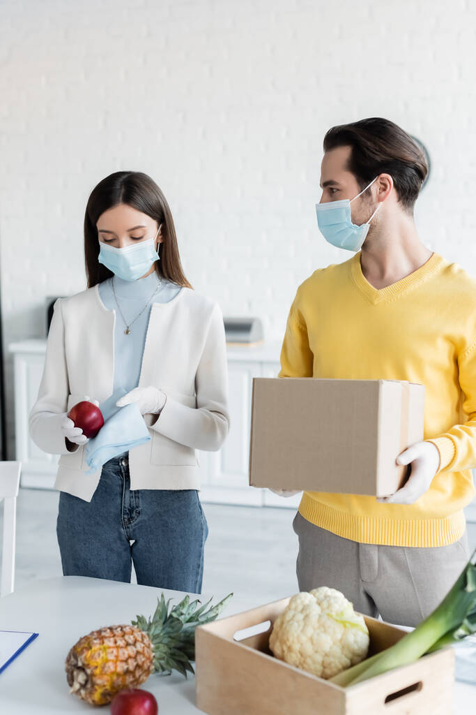 Man in medical mask holding carton box near girlfriend cleaning apple with rag at home  - Φωτογραφία, εικόνα