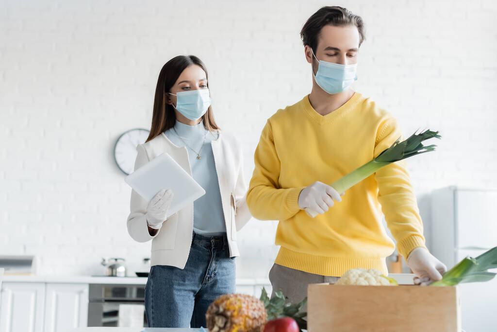Woman in medical mask holding digital tablet near boyfriend in latex gloves taking vegetables from wooden box in kitchen  - Foto, Bild
