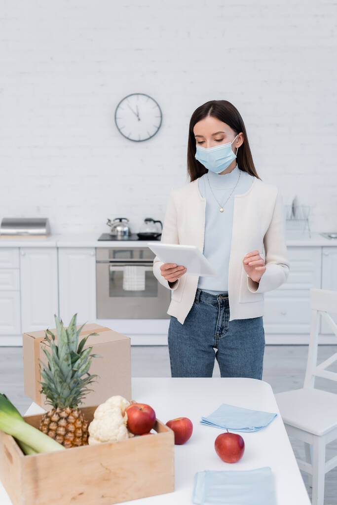 Woman in medical mask holding digital tablet near fresh food and carton box in kitchen  - Foto, imagen