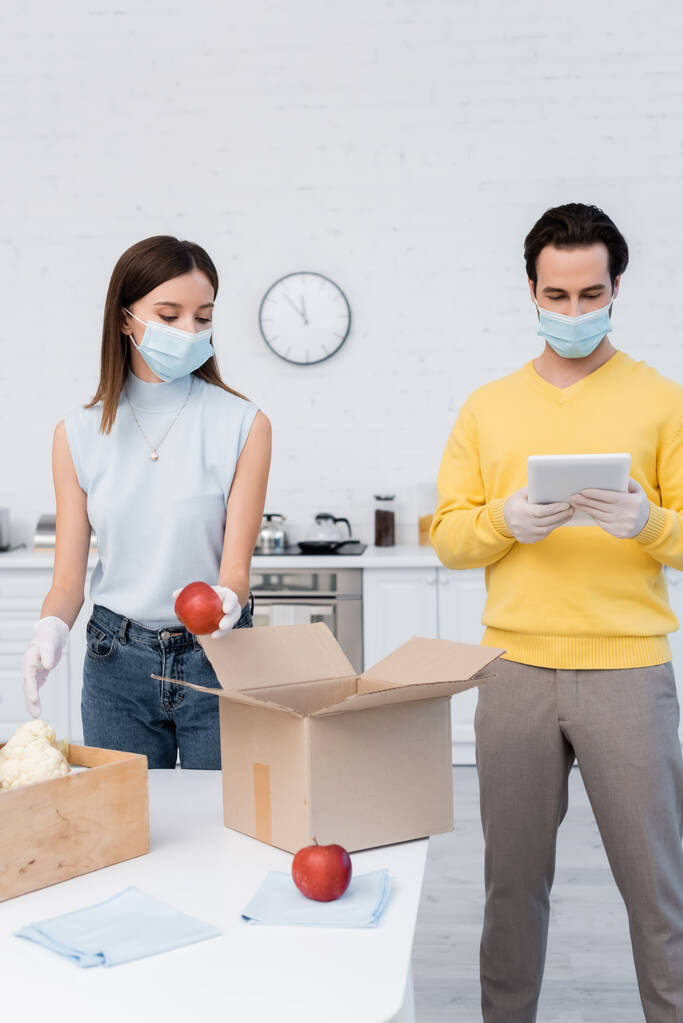 Woman in protective mask and latex gloves holding apple near boxes and boyfriend using digital tablet in kitchen  - 写真・画像