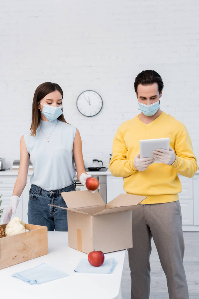Man in latex gloves and medical mask using digital tablet near girlfriend, fresh food and boxes in kitchen  - Photo, image