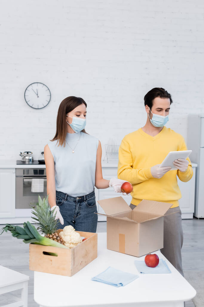 Man in medical mask using digital tablet near girlfriend holding apple and food in boxes in kitchen  - Foto, Bild