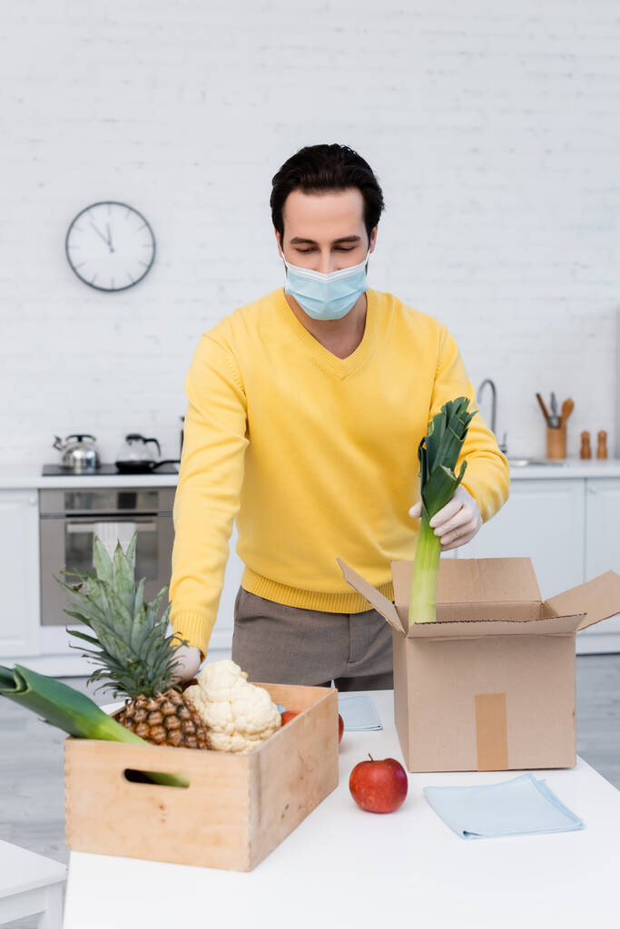 Young man in medical mask holding leek near fresh food and rags at home  - Foto, imagen