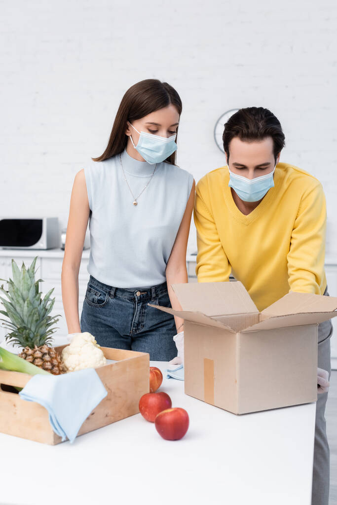 Couple in medical masks looking at carton box near fresh food and rags in kitchen  - Foto, immagini