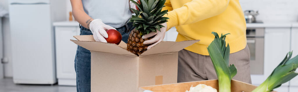 Cropped view of couple in latex gloves taking fruits from box in kitchen, banner  - 写真・画像