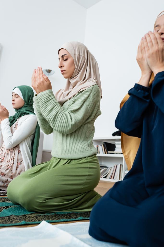 Jovem muçulmano mulher orando no tapete perto asiático mãe e filha em casa  - Foto, Imagem
