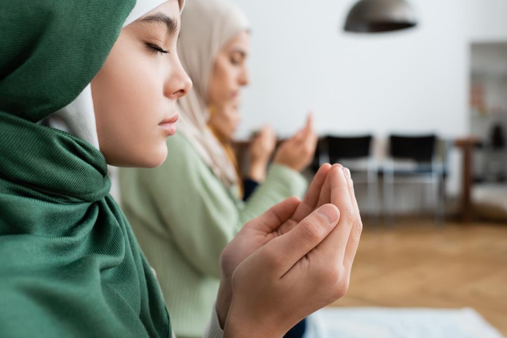 Widok z boku dziewczyny w hidżabie modląc się w pobliżu rozmytej rodziny w domu  - Zdjęcie, obraz