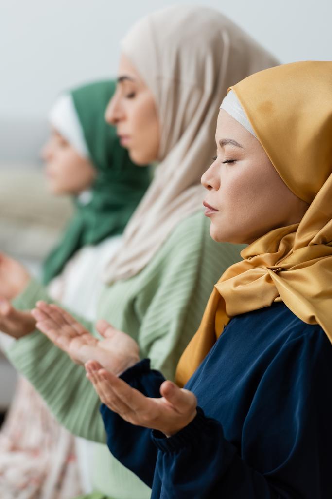 Вид збоку на азіатську жінку в хіджабі, що молиться біля розмитої мусульманської дочки та онуки вдома
  - Фото, зображення