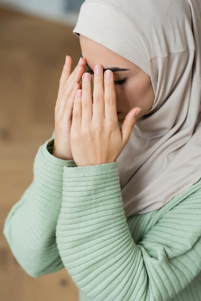 Fiatal arab nő kéz a kézben az arcon, miközben otthon imádkozik  - Fotó, kép