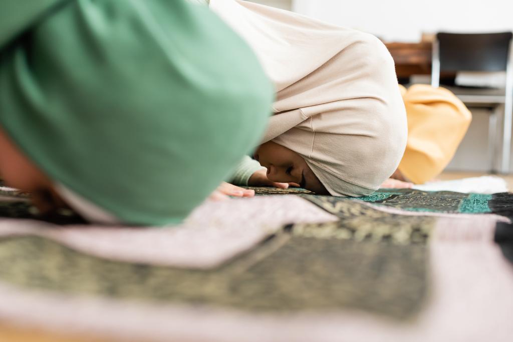Boční pohled muslimské ženy v hidžábu ohýbání na koberec v blízkosti rozmazané dcery, zatímco modlí doma  - Fotografie, Obrázek