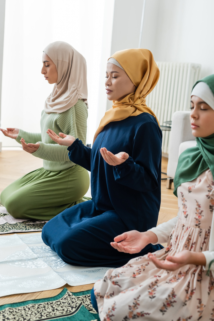 Geleneksel Müslüman kıyafetleri içinde Asyalı bir kadın evde Arap kızı ve torununun yanında dua ediyor.  - Fotoğraf, Görsel