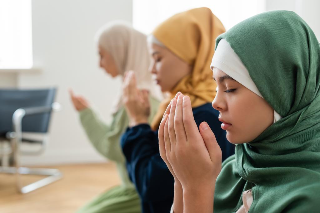 Πλευρική άποψη του κοριτσιού σε hijab κρατώντας τα χέρια κοντά στο πρόσωπο, ενώ προσεύχεται κοντά πολυεθνική οικογένεια  - Φωτογραφία, εικόνα