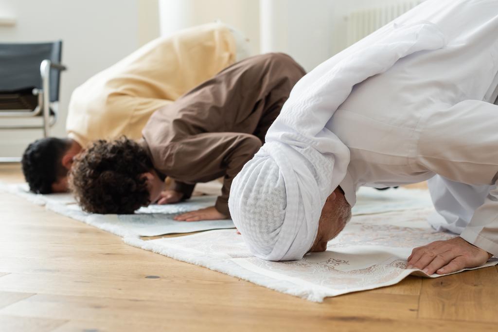 Зрелый человек, склоняющийся на ковре, молясь дома возле арабской семьи  - Фото, изображение