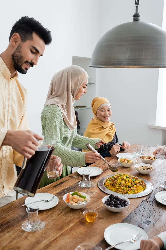 Улыбающаяся арабская женщина, стоящая рядом с едой, муж с чаем и азиатская мама дома  - Фото, изображение
