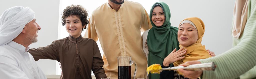 Lächelnde multiethnische muslimische Familie in der Nähe einer Frau mit Pilaf zu Hause, Banner  - Foto, Bild