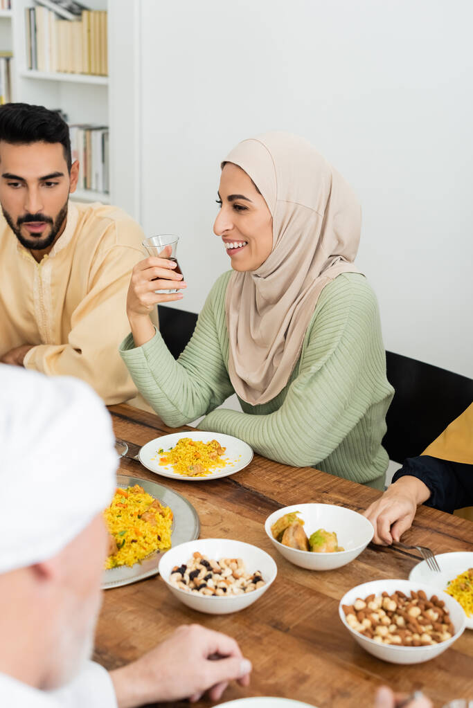 Χαρούμενη Αραβίδα γυναίκα με μαντίλα κρατώντας ένα ποτήρι τσάι κατά τη διάρκεια του δείπνου με την οικογένεια μουσουλμάνων - Φωτογραφία, εικόνα