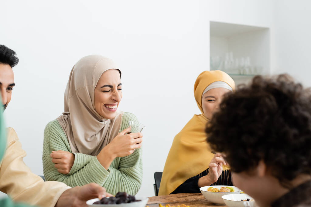 alegre mujer musulmana en hijab riendo durante la cena con la familia multicultural - Foto, imagen
