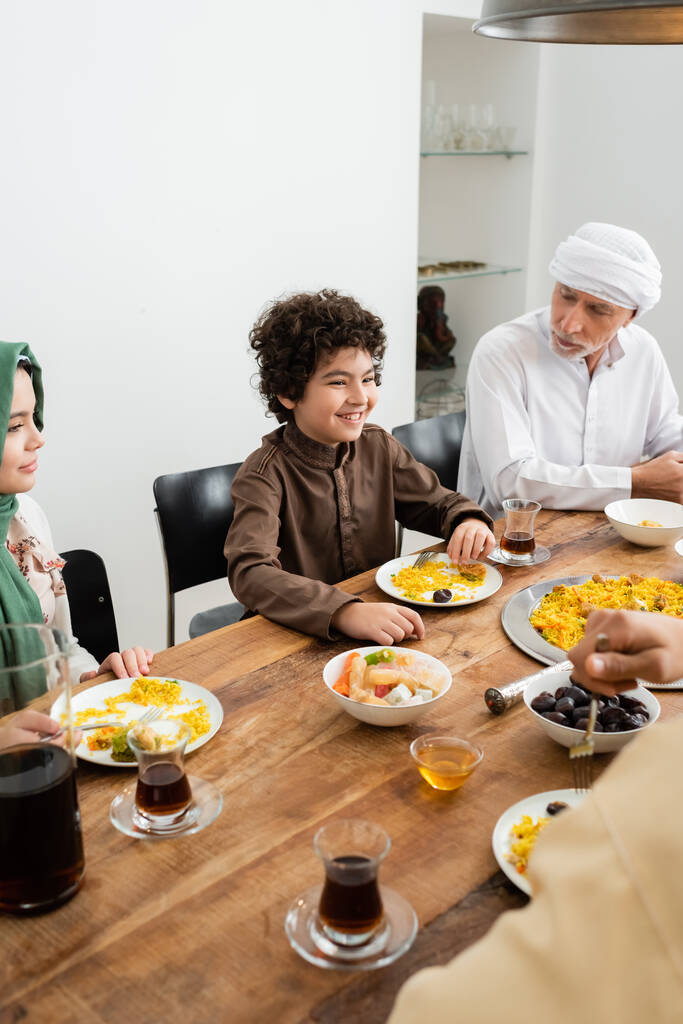 szczęśliwe wielonarodowe dzieci jedzące obiad w pobliżu muzułmańskiego dziadka w domu - Zdjęcie, obraz