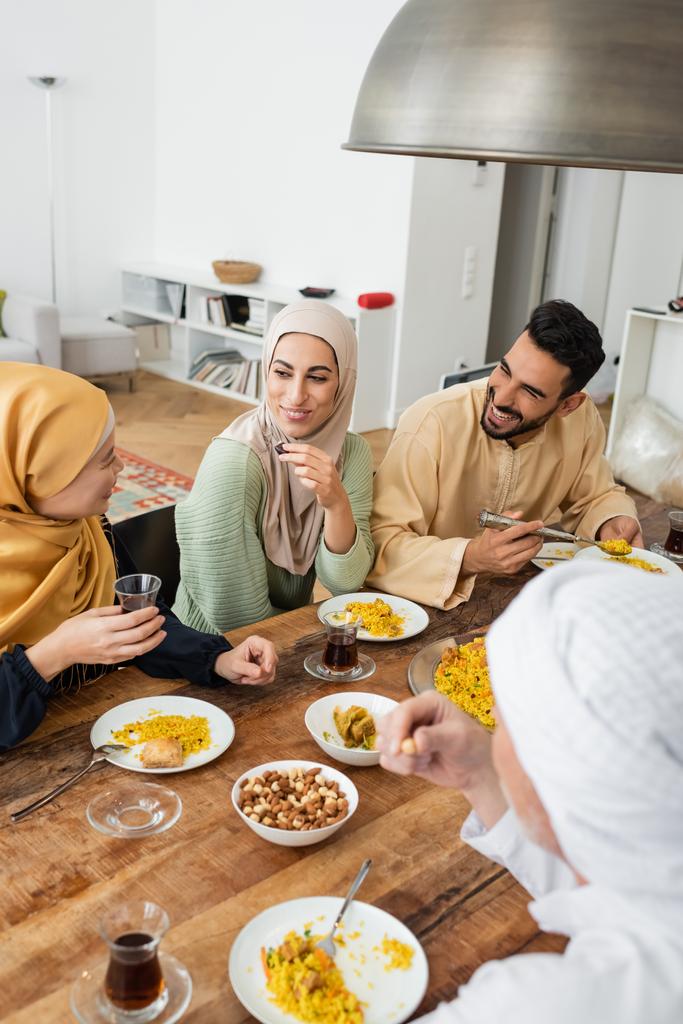mujer asiática madura sosteniendo un vaso de té mientras habla con la familia musulmana durante la cena - Foto, imagen