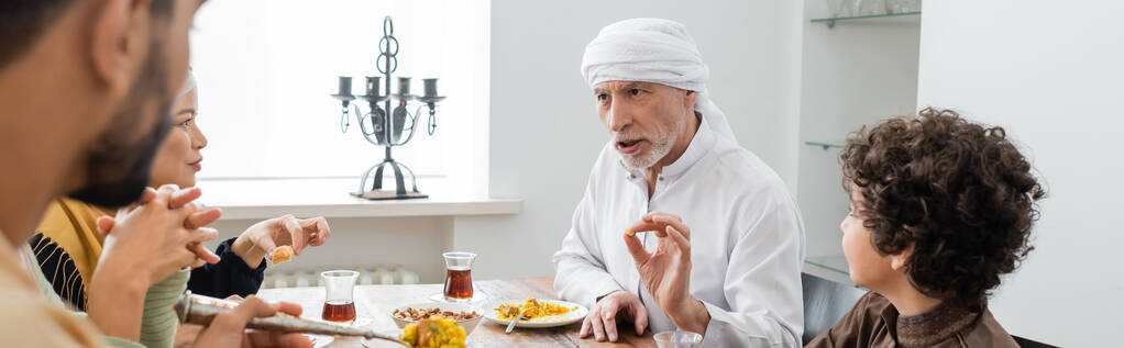 зріла мусульманська людина жестикулює під час розмови під час вечері з мультикультурною сім'єю, банер
 - Фото, зображення
