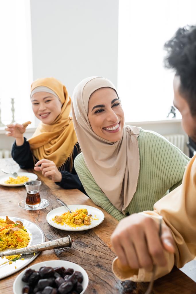 夕食中に夫とアジア系の女性の近くで笑っているヒジャーブの幸せなイスラム教徒の女性 - 写真・画像