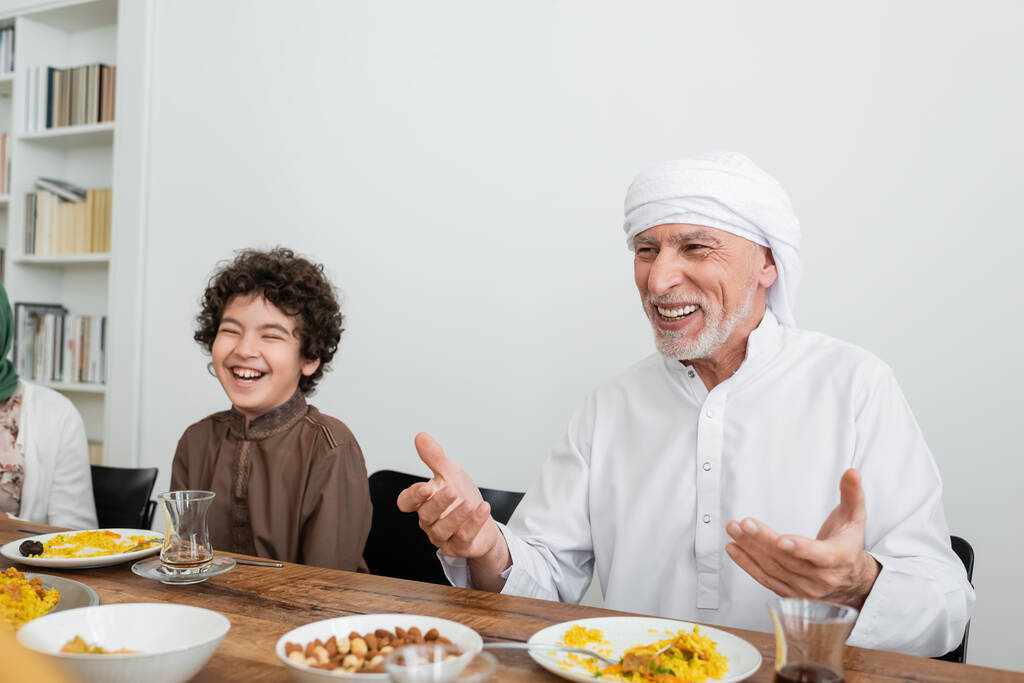 glücklicher muslimischer Mann zeigt beim Familienessen mit Händen in der Nähe des lachenden arabischen Enkels - Foto, Bild