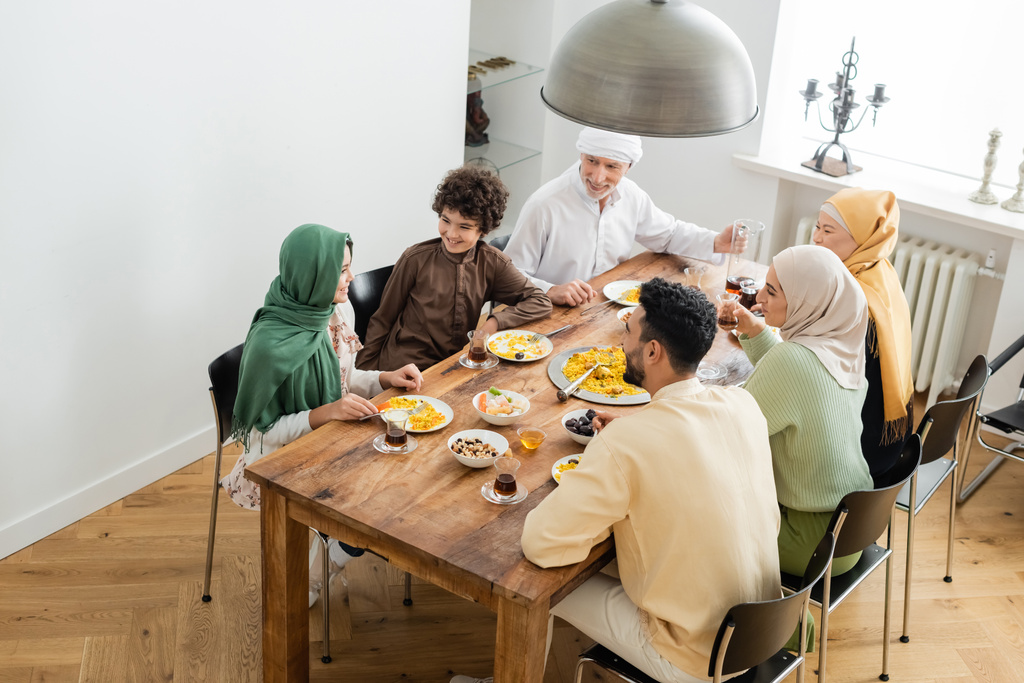 wysoki kąt widzenia międzyrasowej muzułmańskiej rodziny uśmiechniętej podczas kolacji w domu - Zdjęcie, obraz