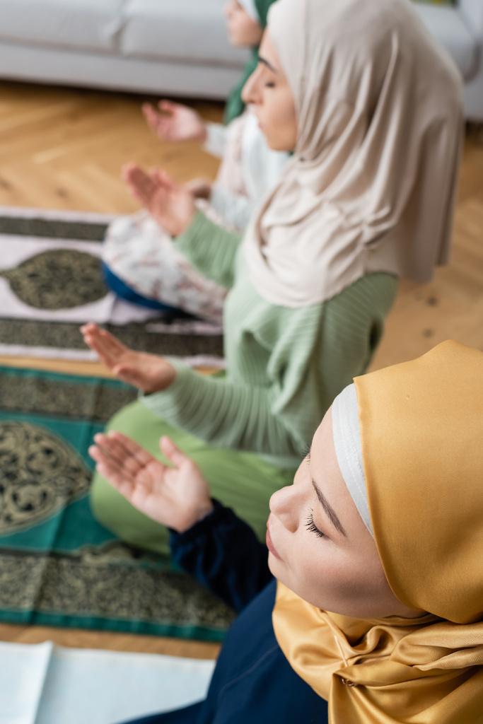 άποψη υψηλής γωνίας της μουσουλμανικής ασιατικής γυναίκας με κλειστά μάτια προσεύχεται κοντά κόρη και εγγονή στο σπίτι - Φωτογραφία, εικόνα