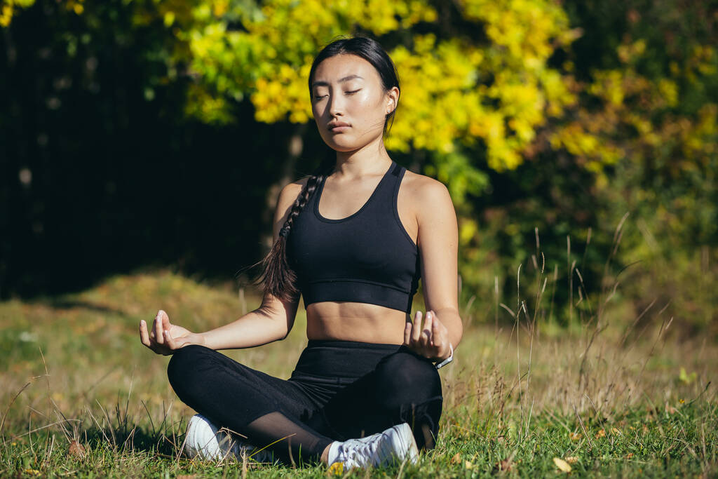 Young beautiful asian woman performs fitness yoga exercises, sitting in lotus position on the grass and meditating in the park - Foto, Bild