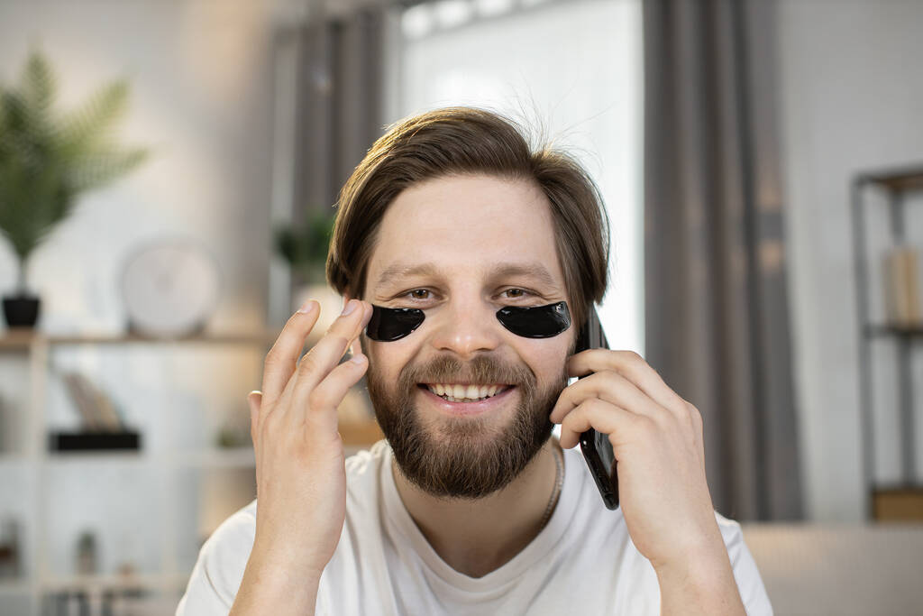 Caucasian man, with black hydrogel patches under eyes, sitting at home and talking on the phone - Photo, Image