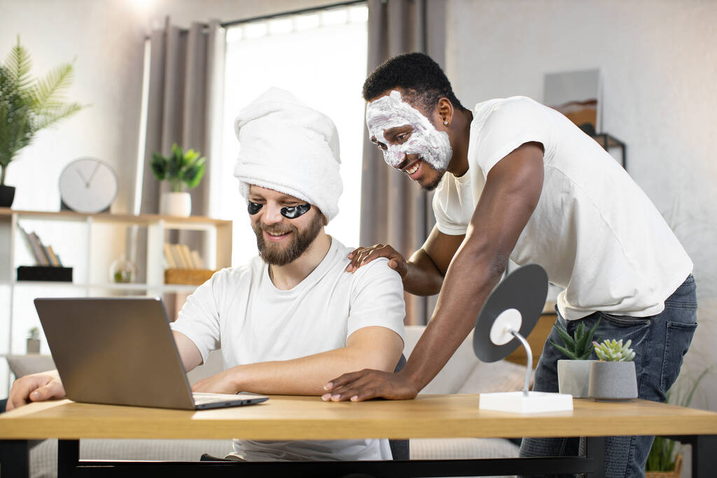 Πολυφυλετικές δύο άνδρες φίλοι με μπαλώματα ματιών και μάσκα προσώπου γελώντας βλέποντας αστεία βίντεο - Φωτογραφία, εικόνα