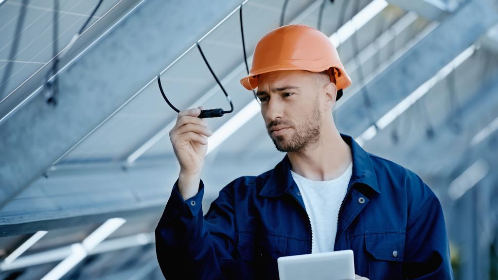 engineer in hardhat checking wire of solar panels while holding digital tablet  - Фото, изображение