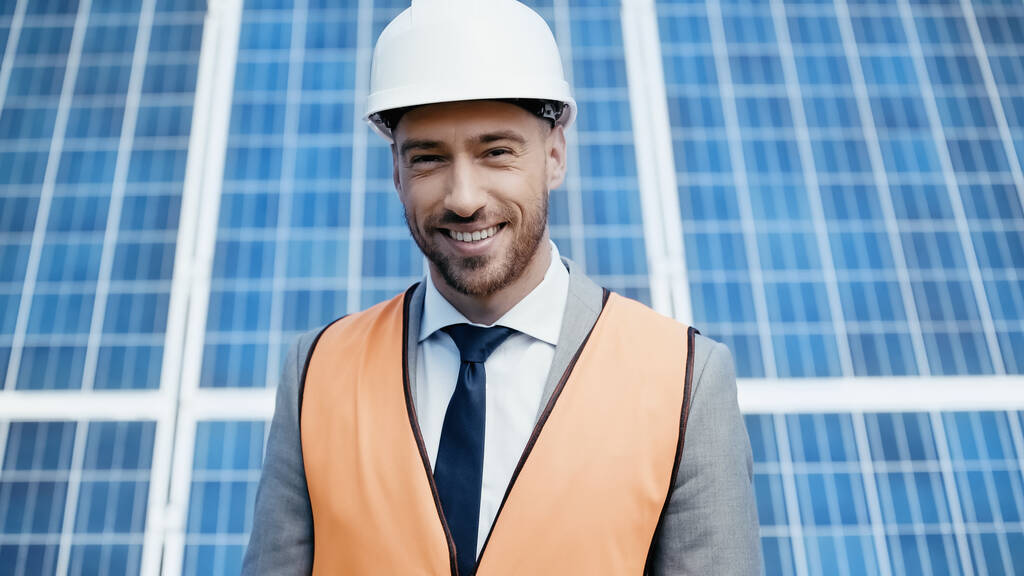 alegre hombre de negocios en hardhat y chaleco de seguridad cerca de las baterías solares - Foto, Imagen