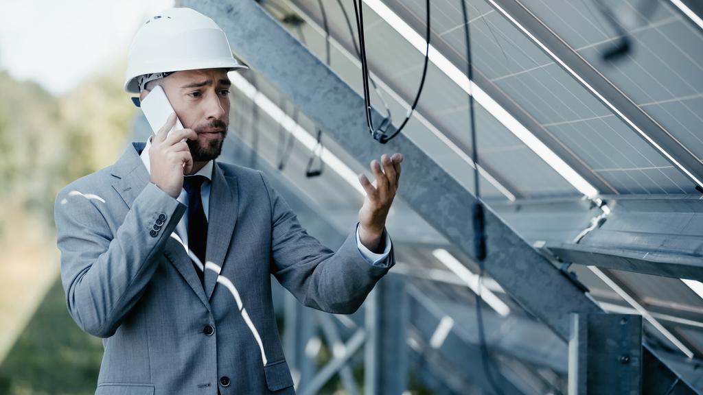 empresario disgustado mirando alambre colgante mientras habla en el teléfono inteligente cerca de paneles solares - Foto, imagen