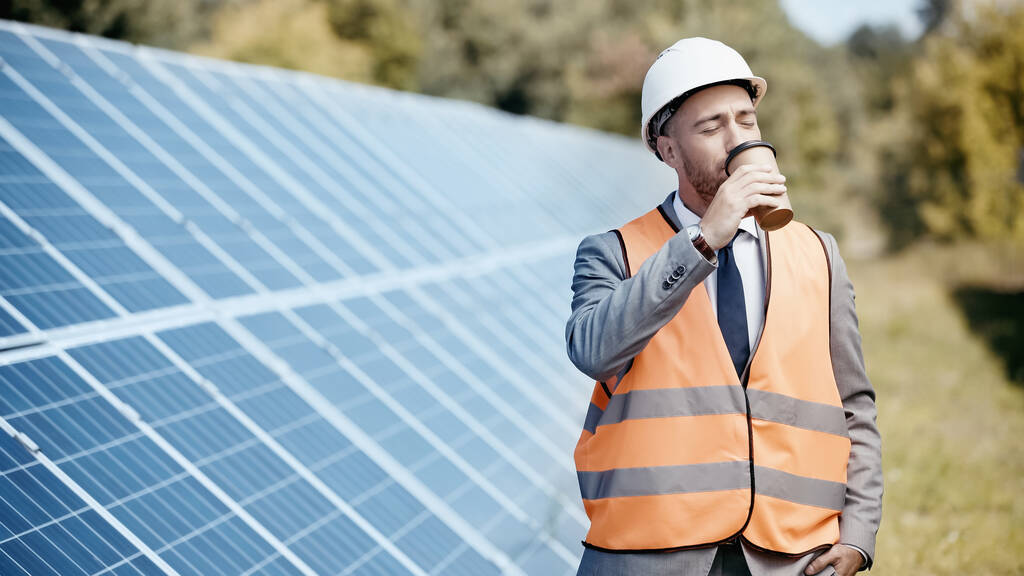 businessman in hardhat drinking coffee to go near solar batteries outside - Photo, image