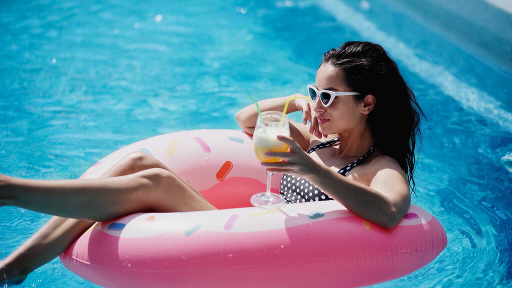 fröhliche Frau mit Sonnenbrille hält Glas mit Cocktail in der Hand und schwimmt auf aufblasbarem Ring im Pool - Foto, Bild