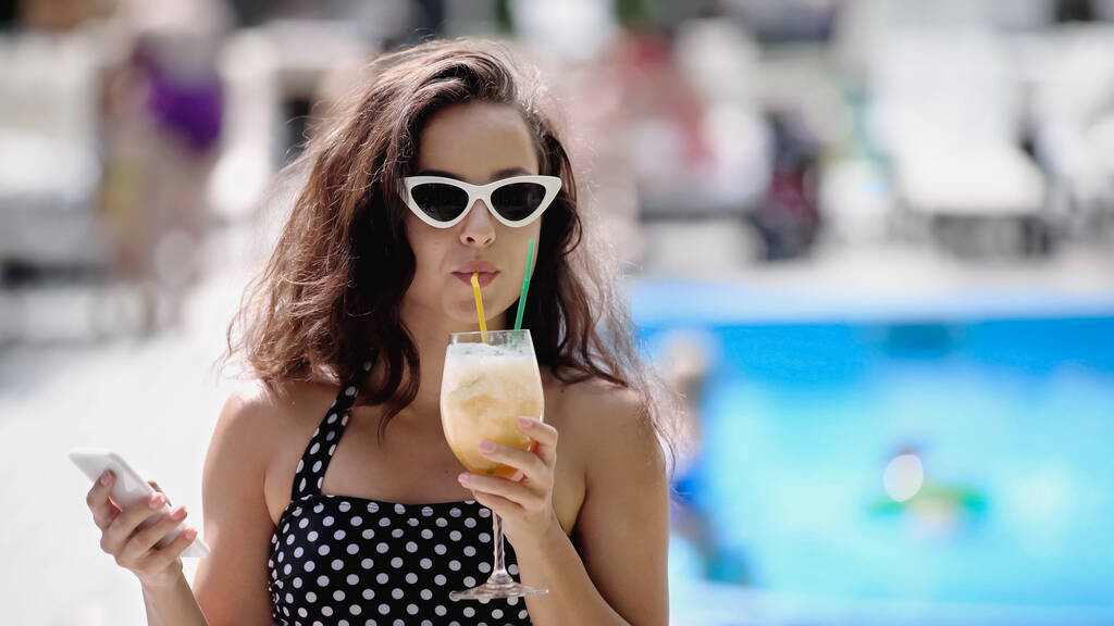 brünette Frau in Sonnenbrille und Badeanzug trinkt Cocktail und hält Smartphone in der Hand - Foto, Bild