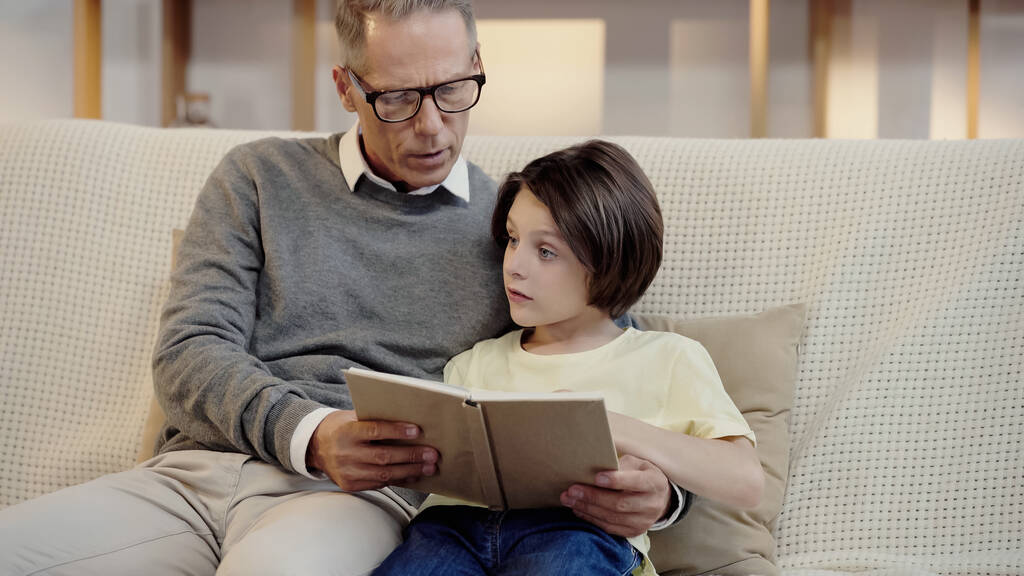 grandfather in glasses reading book to grandson at home - Photo, image