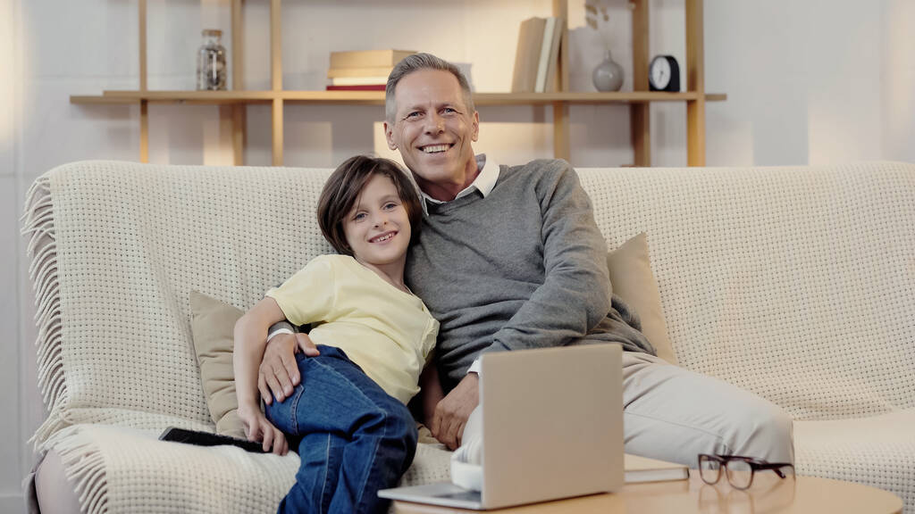 dziadek w średnim wieku uśmiechnięty ze szczęśliwym wnukiem podczas oglądania filmu w domu - Zdjęcie, obraz